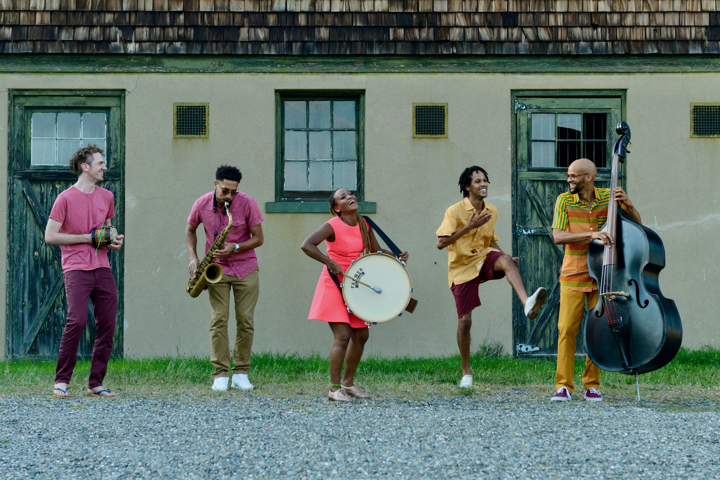Members of Music From The Sole stand in a line jamming in front of a farmhouse building, wearing colorful yellow, pink, and orange clothes.